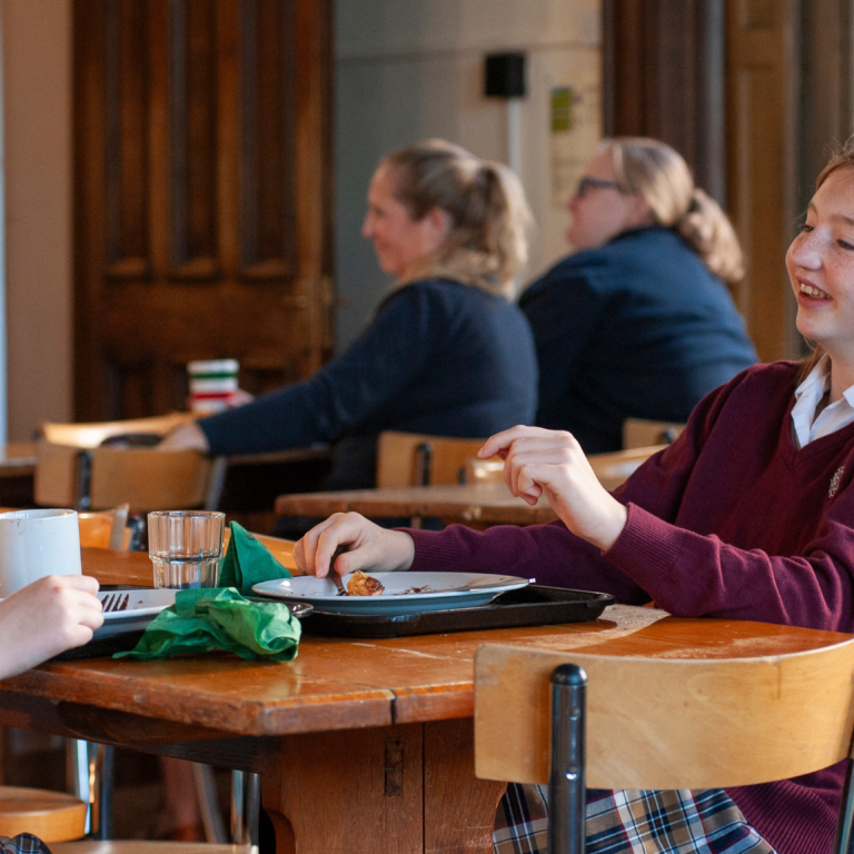 Two young women sit opposite each other at a cafeteria table, smiling and chatting over plates and mugs.