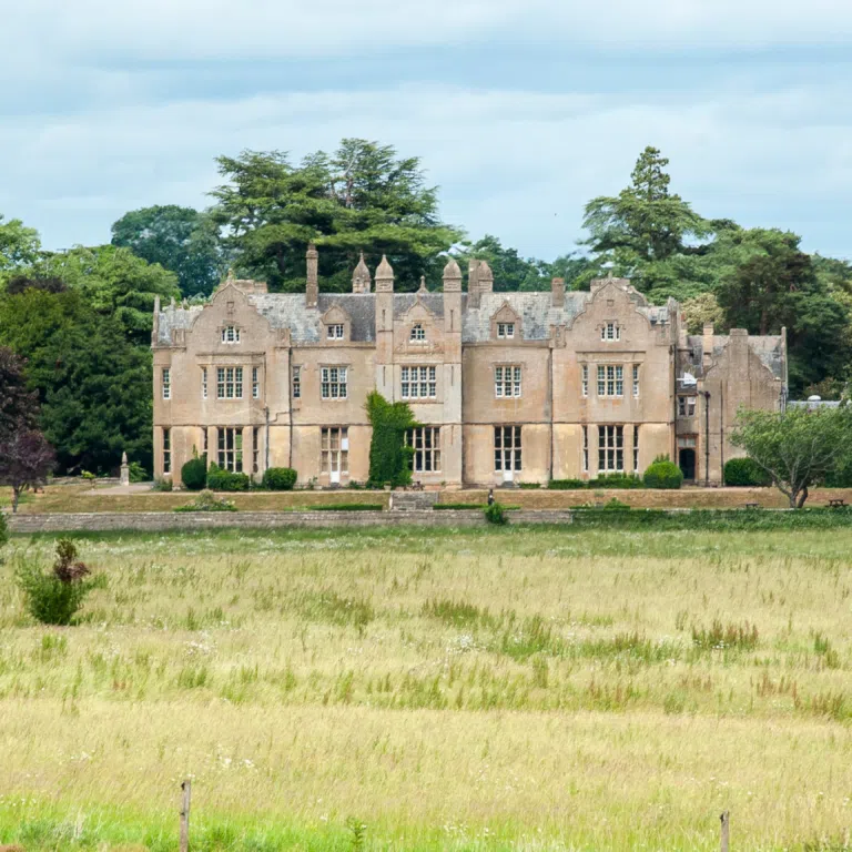 Stone manor house with ornate gables in a green park, surrounded by trees under a cloudy sky.