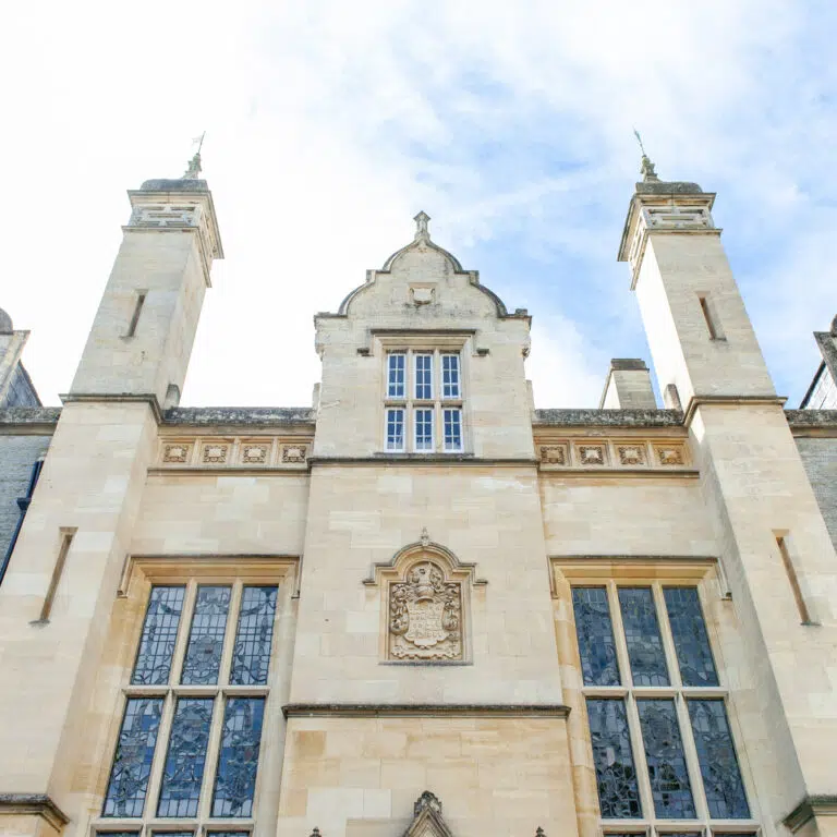 Front fa&ccedil;ade of a historic stone building with tall towers and decorative windows against a blue sky.