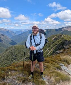Hiker standing on a rocky mountain ledge with backpack and trekking poles, overlooking forested valleys under a blue sky with clouds.