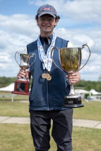 Young man in a blue cap with the Union Jack, holding two trophies and wearing medals outdoors.