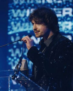Man with wavy dark hair speaks into a handheld microphone on stage, wearing a sparkly black jacket with a clear podium nearby.