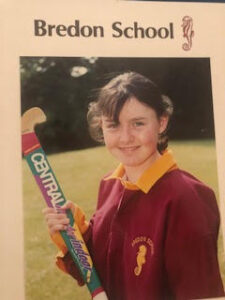 Portrait of a smiling girl in a maroon Bredon School uniform with a yellow collar, outdoors on a field, holding a cricket bat with the school crest visible.
