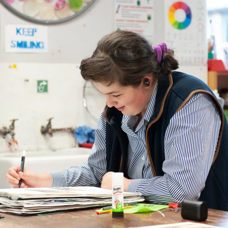 Person wearing a blue striped shirt and purple hair tie draws with a black marker at a cluttered desk, papers scattered and a Pritt glue stick nearby.