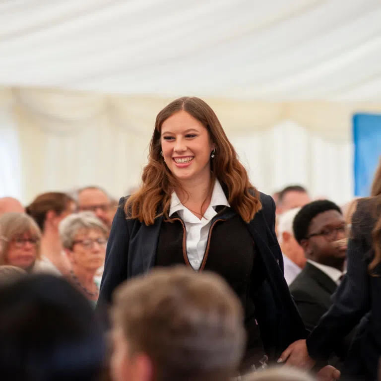 Smiling young woman with long wavy hair in a navy blazer and white shirt, standing in a crowded indoor event.