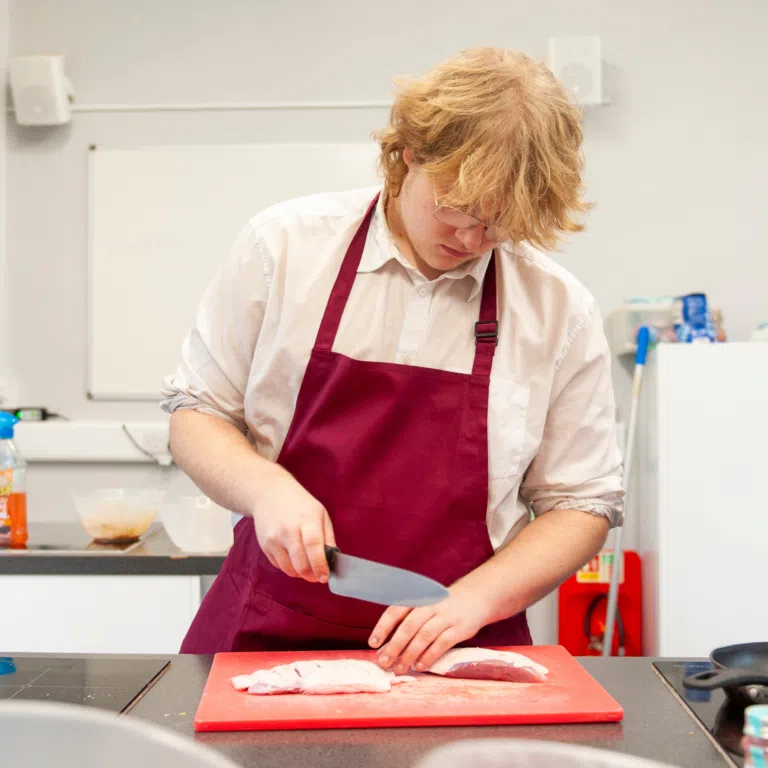 Chef or cook in a white shirt and maroon apron slicing meat on a red cutting board in a kitchen