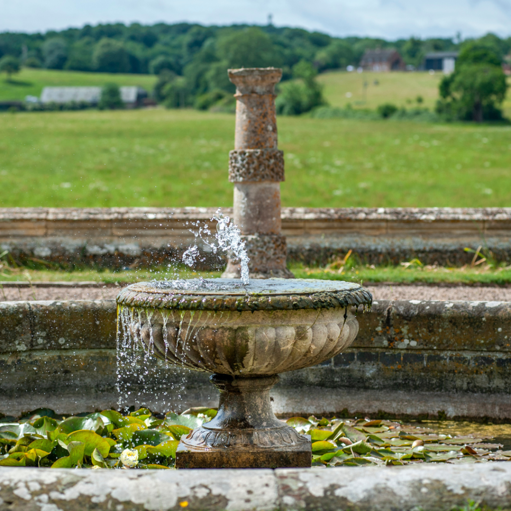 Decorative stone garden fountain with water spraying from the rim, surrounded by lily pads and a rural field in the background
